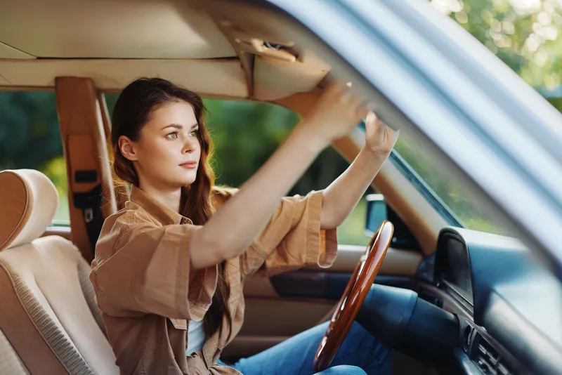 Young woman adjusting the mirror in a classic car, showcasing a casual outfit with a joyful expression, surrounded by a vibrant outdoor setting.
