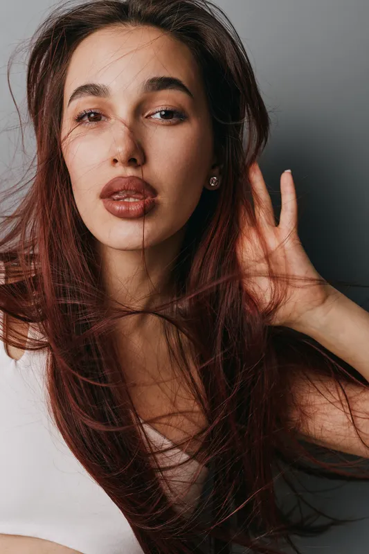 Woman with long hair posing in studio with hand near ear and natural makeup, looking at camera with confident expression on grey background closeup portrait.