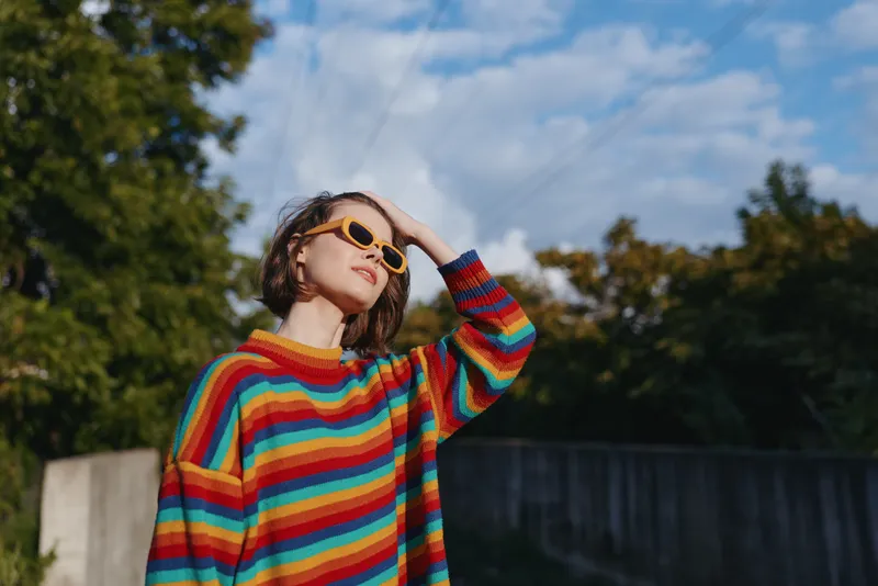 Woman in her 20s wearing a colorful striped sweater and sunglasses outdoors, smiling and looking up with hand on head, casual retro style in a sunny urban park lifestyle scene.