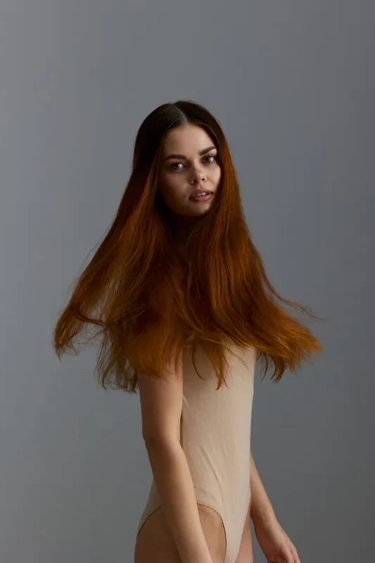 Beauty woman with long flowing brown hair and natural makeup wearing beige bodysuit against neutral gray background, showcasing fashion style and graceful poise.