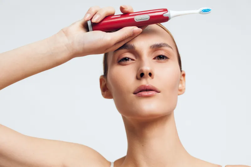 woman beauty skincare shaving grooming hygiene tool closeup, young woman holds red trimmer to forehead, clean white background, minimal spa routine, calm focused face, skin care ritual
