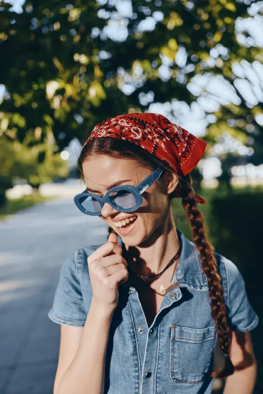 smiling woman with braided hair wearing blue sunglasses and red bandana outdoors in sunny park with green trees and casual denim outfit