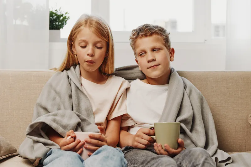 Two children wrapped in gray blankets sit on a couch drinking warm beverages, showing a cozy and relaxed indoor moment during cold weather or family time.