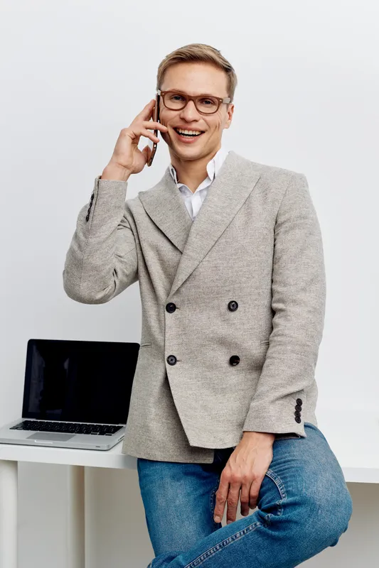 Smiling man in business casual attire talking on mobile phone indoors with laptop on white table behind him, expressing happiness and confidence in a modern office environment. People lifestyle concep