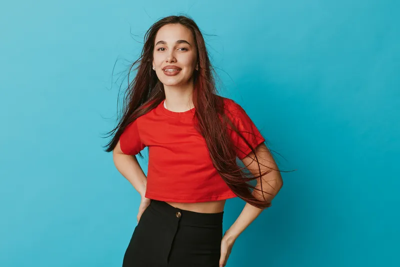 Young woman with long hair wearing red shirt and black pants posing confidently against blue background. Casual fashion, smiling female model with natural makeup and relaxed posture.