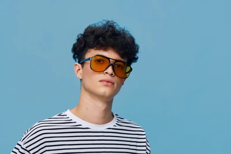 Young man with curly hair wearing yellow sunglasses and striped shirt posing confidently against a blue background in casual style.