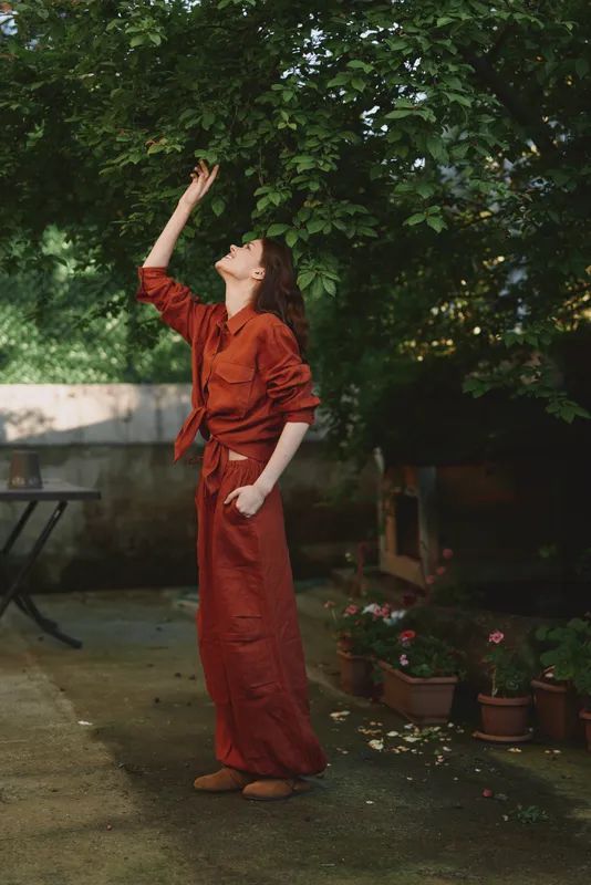 A woman in a rust dress reaches toward the leaves in a shaded garden, capturing a moment of curiosity and connection with nature, her calm stance and soft light creating a contemplative mood