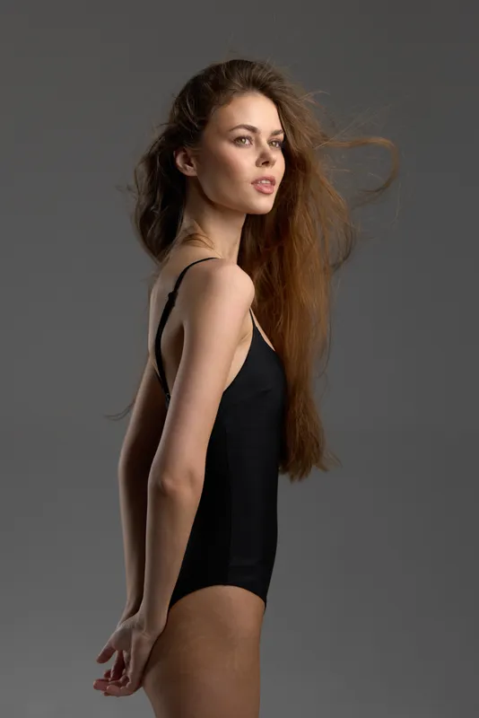 Young woman in black bodysuit standing sideways with flowing hair and neutral expression against gray background
