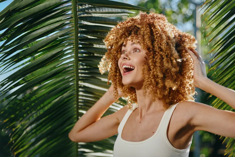smiling woman with curly hair wearing white tank top enjoys sunny tropical background with green palm leaves, natural light, and joyful summer vibe.