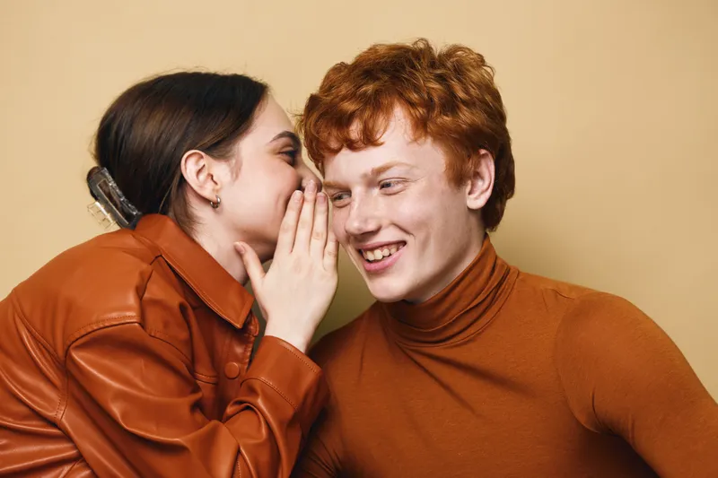 Young woman whispers secret to smiling redhead man in casual clothes, closeup with indoor studio background, interaction and friendship between cheerful people.
