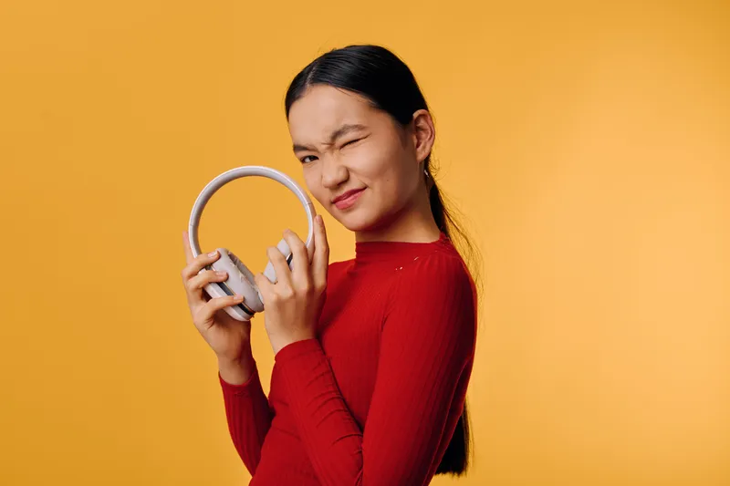 Woman in red sweater holding headphones with playful wink and smile against yellow background, casual portrait with relaxed mood and bright studio lighting for lifestyle concept.