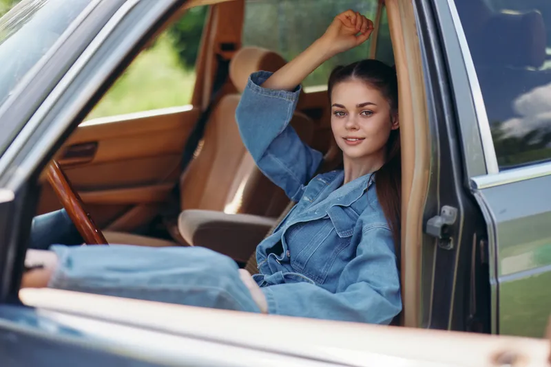 Young female in denim outfit relaxing inside a car, showcasing casual style and confidence in a vibrant, green outdoor setting.