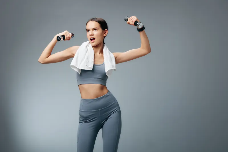 Fitness woman in athletic wear lifts dumbbells with a towel over her shoulders, dynamic workout pose, intense focus and energy showcasing strength, sport and exercise.