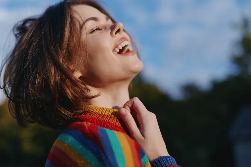 Young adult woman (mid 20s) smiling and laughing in a colorful rainbow sweater, short hair portrait outdoors at golden hour, joyful mood for lifestyle and fashion travel.