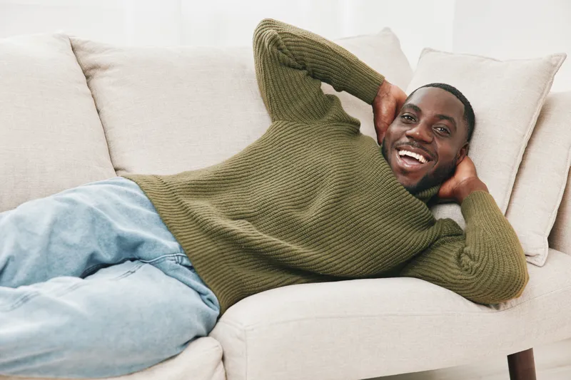 Young Black man in a cozy green sweater relaxing on a couch, exuding happiness, with a bright background and soft colors Lifestyle concept of comfort and joy