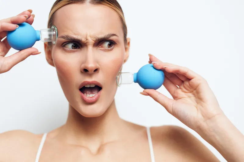 Woman, facial, suction, massage, skincare, close up, blue cupping tool, surprised expression, indoor, white background, head and shoulders, removing puffiness