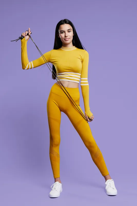 Fit woman sport fitness wearing bright yellow activewear holds jump rope, standing confidently against a purple background with athletic posture and white sneakers.