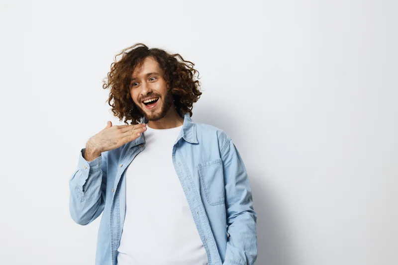 Happy young man with curly hair in a blue shirt expressing joy, standing against a white background, perfect for lifestyle and emotion themes