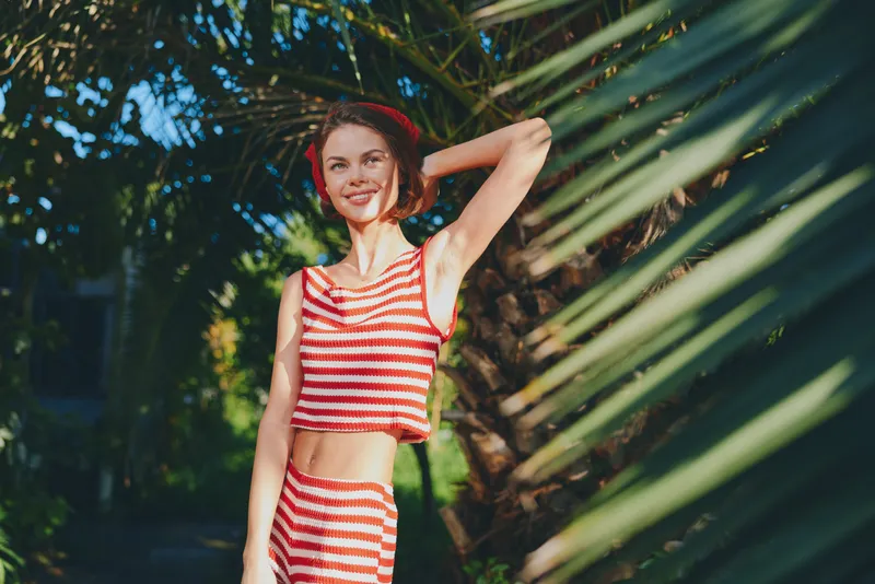 A cheerful young woman stands outdoors in a sunny garden, wearing a striped red and white crop top and shorts. She smiles warmly and poses confidently among lush greenery.