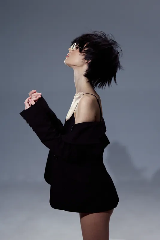 Young woman in black outfit posing with closed eyes and tousled hair against gray background, showing stylish and moody expression.