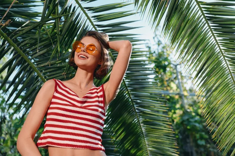 A cheerful woman in a red and white striped tank top wears sunglasses and enjoys a sunny outdoor moment among lush palm trees, projecting a confident and carefree vibe in a bright tropical setting.