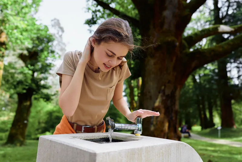 Woman girl drinking water from a public fountain in a park, outdoor summer scene with trees and grass, casual clothing, refreshing hydration and candid moment on a warm day.