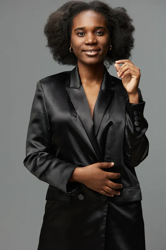 Confident Black woman in a stylish satin blazer smiling with natural curly hair against a soft gray background, conveying empowerment and professionalism