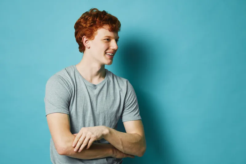 Young man with red hair smiling and looking to side wearing casual grey t shirt against blue background in studio. Portrait of happy male with crossed arms and relaxed posture.