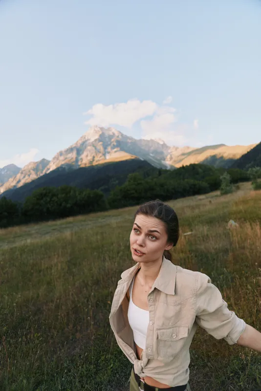 Woman in casual attire stands in a meadow with distant mountains, showcasing an outdoorsy lifestyle, calm nature, and exploration under soft natural light. Warm glow.