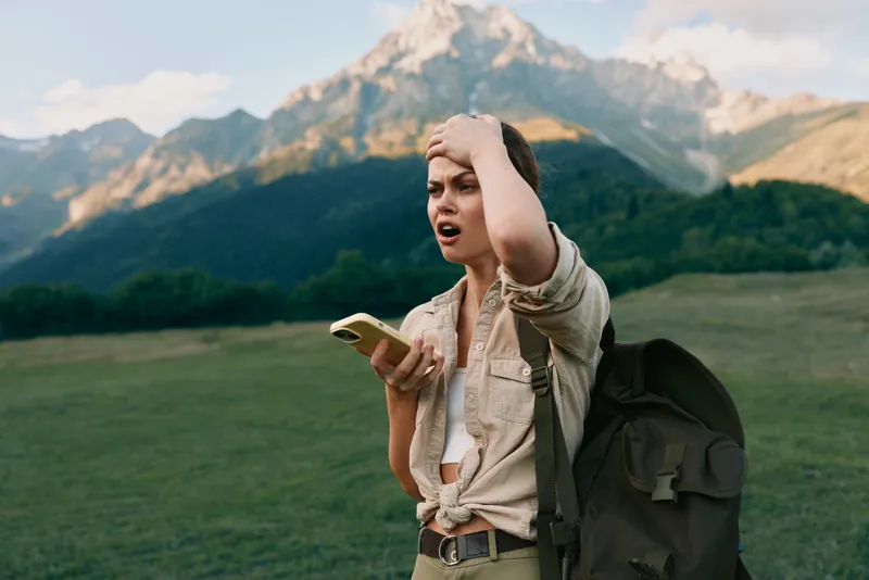 Casual woman outdoors with a backpack holding a phone, surprised expression, standing on a grassy field with mountains in the background, travel lifestyle moment, spontaneous adventure