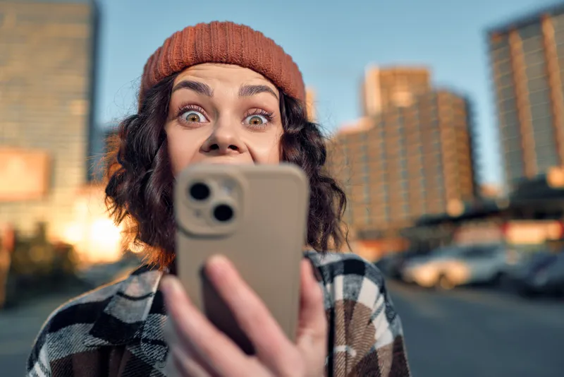 Woman with phone taking a selfie in urban street, surprised expression and beanie, candid lifestyle shot with golden hour glow, authenticity and emotional storytelling of mindful living.
