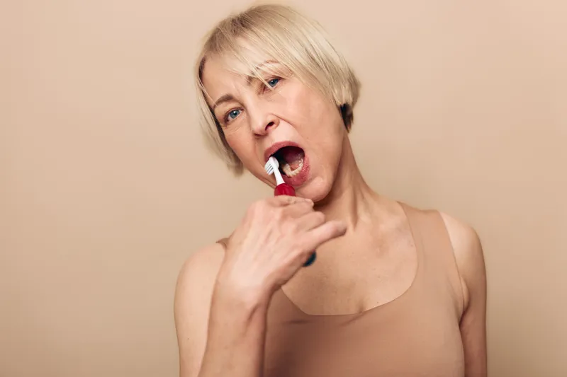 Middle aged woman with short blonde hair brushes teeth using white toothbrush, wearing beige tank top. Neutral background emphasizes oral hygiene, daily routine, and personal care concept.