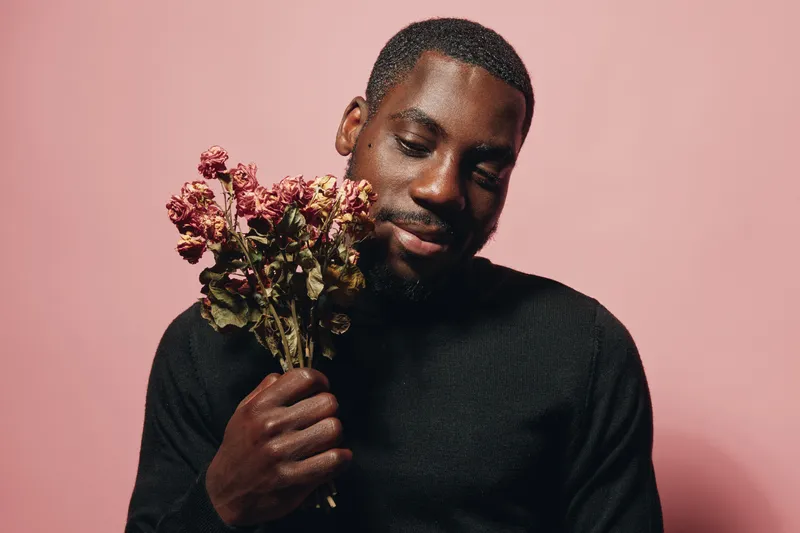 African man holding dried flowers with closed eyes in creative conceptual visual style on pink background. Portrait of black male with beard wearing black sweater in studio.