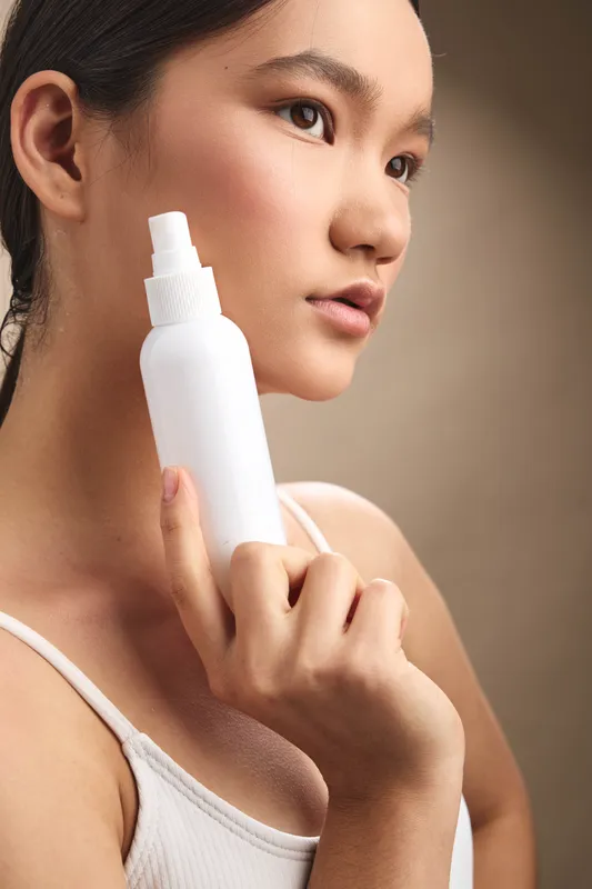 Asian woman applies skincare spray in a calm home setting, her clear skin and focused gaze conveying care and wellness.