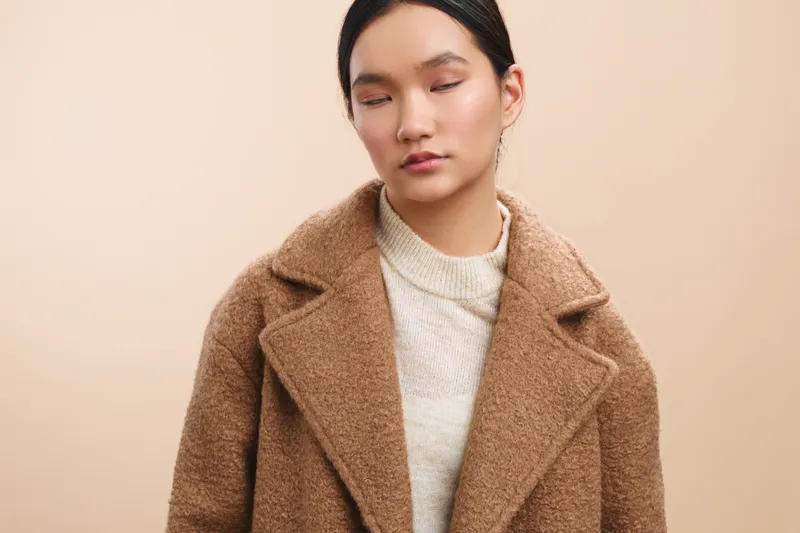 Asian woman in a warm coat stands in a soft studio setting, exuding calm confidence and poise. Her neutral expression and gentle light create a serene, contemporary mood.