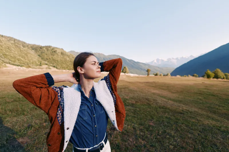 Woman outdoors mountains meadow relaxation pose, hands behind head, wearing jacket with sherpa and denim shirt; portrait of confident solo traveler enjoying scenic valley and clear sky.