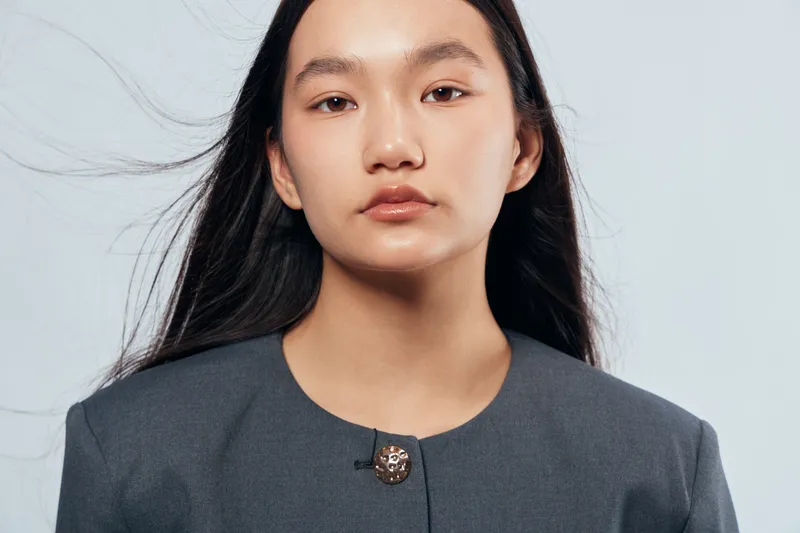 Portrait of young woman with long black hair wearing formal jacket standing against light background. Serious expression with natural makeup and wind blowing strands of hair for style.
