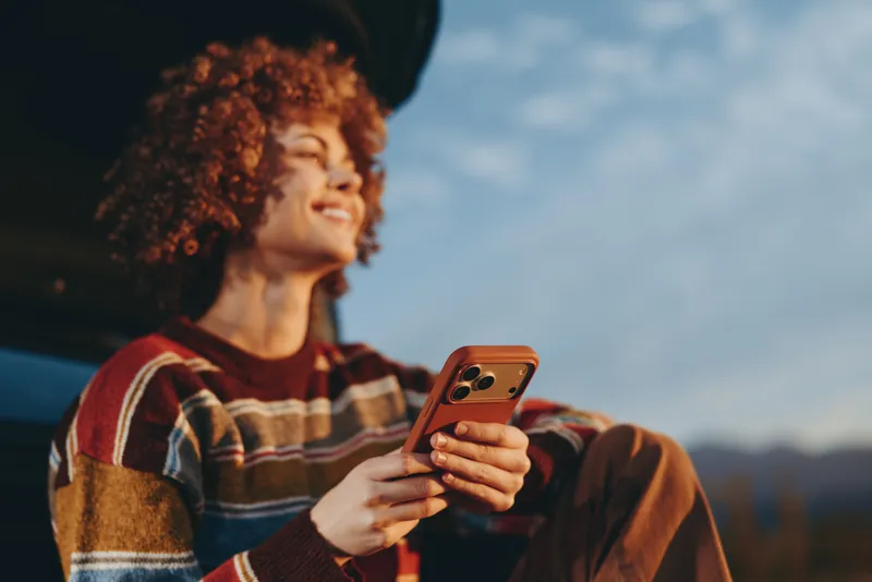Curly haired woman lifestyle outdoors, smiling while holding smartphone, relaxed sitting near car, warm sunset light, soft breeze, mountain landscape in background, cozy scarf, travel pause