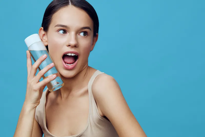 Young woman expressing excitement while holding a skincare product against a blue background, showcasing healthy skincare and beauty regimens with vibrant summer vibes