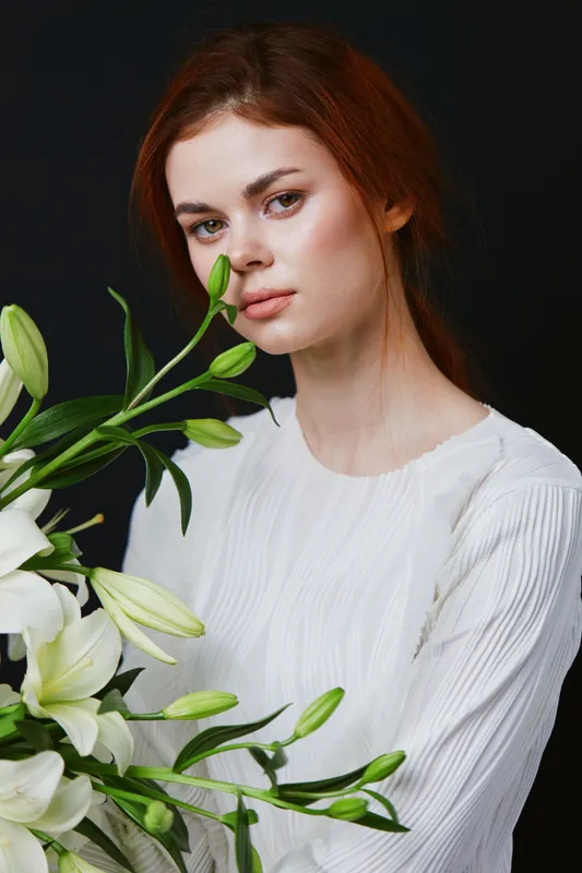 Beautiful young model with long red hair holding white lilies against a dark background, exuding serenity and elegance in a soft white dress
