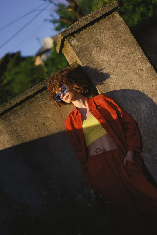 A stylish woman in a red jacket and yellow top leans against a concrete wall, wearing blue sunglasses. The bright urban setting conveys a casual lifestyle and confident attitude
