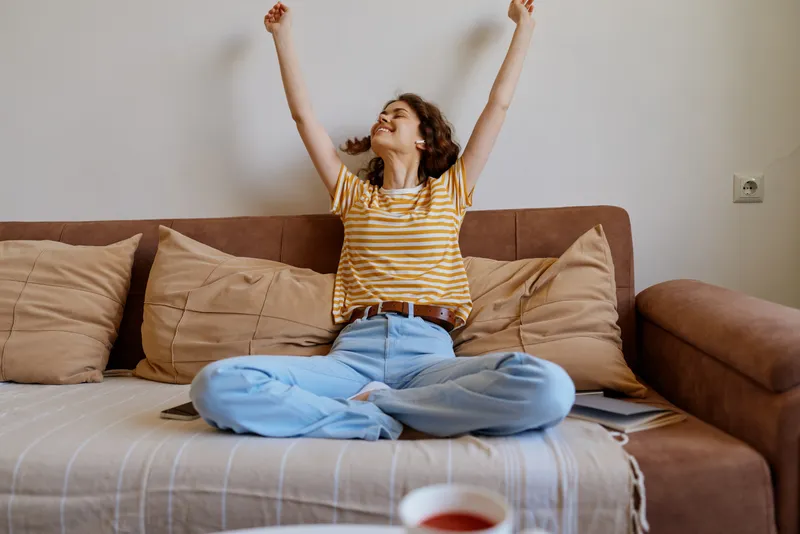 Cheerful woman indoors, sitting crosslegged on a sofa, arms raised in celebration, casual striped shirt and jeans, cozy living room, soft natural light, relaxed lifestyle moment, home comfort