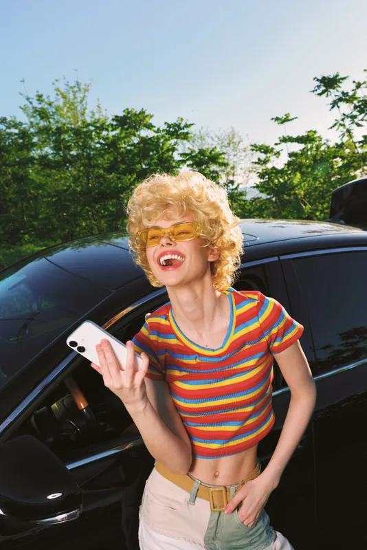 Cheerful woman outdoors, laughing with sunglasses, rainbow striped top, beside a car, sunlit moment, relaxed posture, warm breeze, vibrant colors, casual fashion lifestyle
