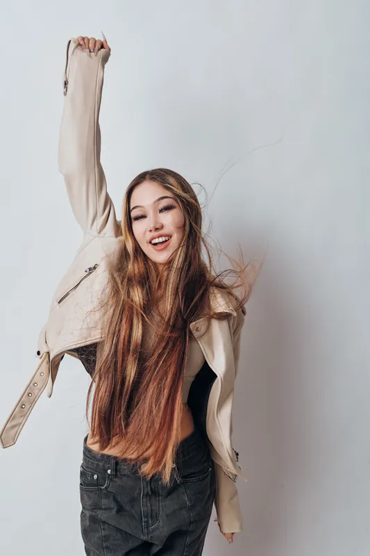 Young woman with long hair wearing beige jacket and black jeans smiling joyfully with raised arm against white background in studio for lifestyle and fashion concept.