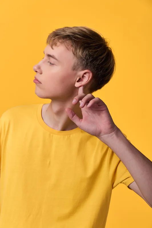 Boy with yellow shirt in profile, ear near a hand as he listens, bright studio backdrop, minimal style, casual mood and modern fashion vibe, emphasizing calm concentration and clean composition