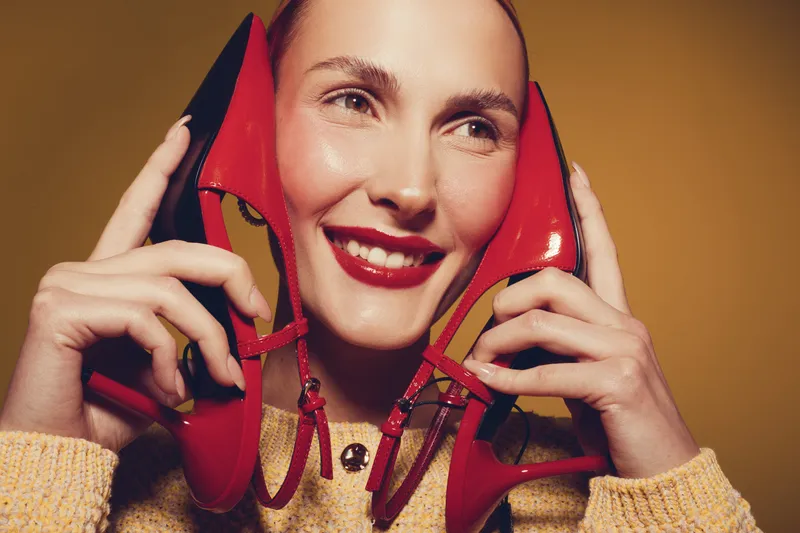 Smiling woman with red lipstick holds red high heel shoes near face. Close-up portrait on warm brown background. Concept of fashion, beauty, and joyful lifestyle with stylish accessories.