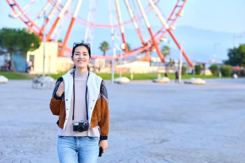 Woman traveler with a camera stands in an outdoor amusement park near a ferris wheel, smiling in a casual jacket and jeans, showcasing travel, leisure and photography in a bright daytime setting