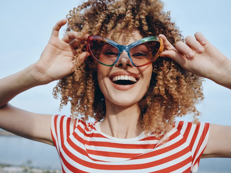 Joyful portrait of a young woman with curly hair wearing oversized colorful sunglasses, smiling broadly toward the camera under a bright clear sky.