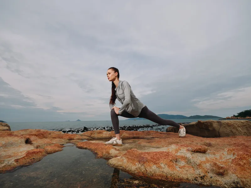 Female model stretching on rocky beach, wearing athletic wear, showcasing determination and fitness in an outdoor setting, with ocean backdrop and cloudy sky