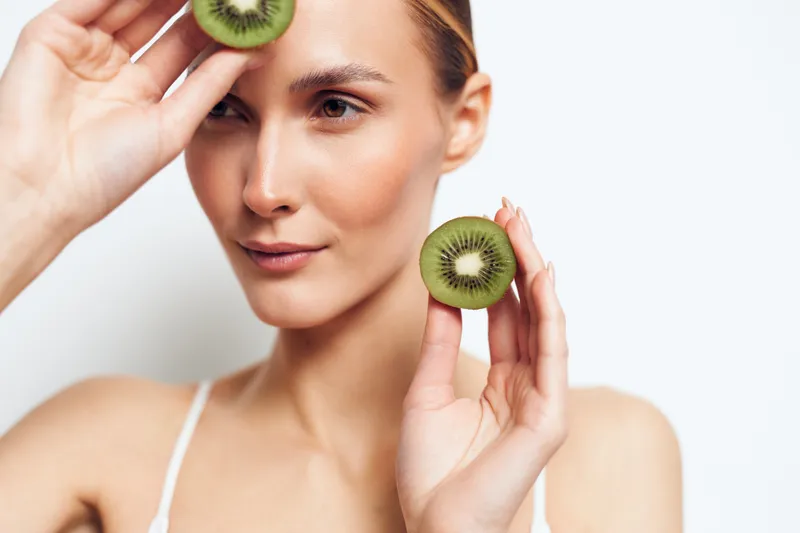 Woman skincare closeup with kiwi slices. She holds a green fruit round on her cheek and near her eye. Soft studio light, clean background, beauty fresh wellness selfcare